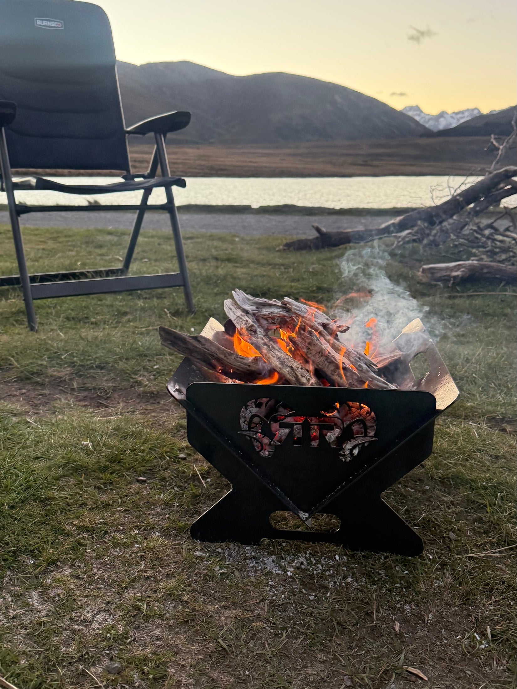 Fire pit with burning logs near a lake and mountains at sunset.